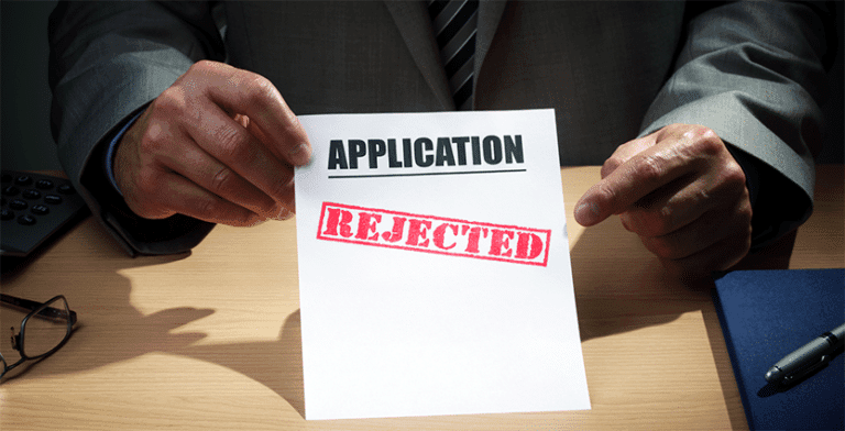 The image shows a gentleman holding a sign on a table that reads, "Application has been rejected." This reflects banks' denials of loans, mortgages, or insurance claims.