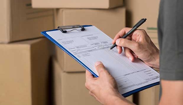 The image shows a warehouse manager signing the dispatch sheet. A close-up of the manager's hand checking the delivery order is visible. A young freight manager is reviewing the invoice alongside a cardboard box parcel.