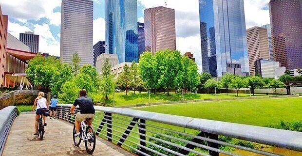 Bicyclists cross wooden bridge in Buffalo Bayou Park, with a beautiful view of downtown Houston