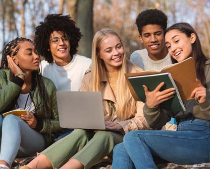 The image shows diverse students collaborating on a group project while studying together in a university or school park.