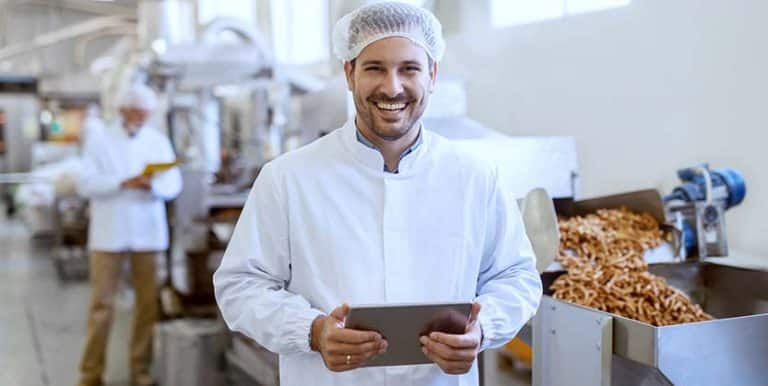 The image shows a young, smiling manager in a sterile uniform holding a tablet and looking at the camera while standing in a food processing and packaging factory.