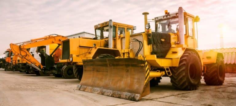 The image shows two heavy-wheeled tractors, one excavator, and other construction machinery in the morning sun.