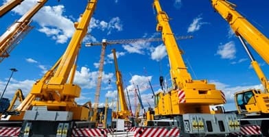 The image shows mobile construction cranes with yellow telescopic arms and large tower cranes on a sunny day. White clouds and a deep blue sky in the background represent heavy industry.