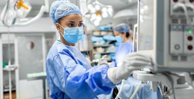 The image shows an anesthetist working in the operating theatre, wearing protective gear, checking monitors while sedating a patient before a surgical procedure in the hospital.