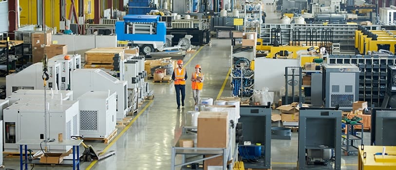 The image depicts a full-length portrait of a businessman, wearing a hard hat, walking through a production workshop alongside a female factory employee.