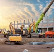 The image depicts multicolored heavy industrial machinery equipment at a construction site parking area against a warehouse building, highlighting city infrastructure development.
