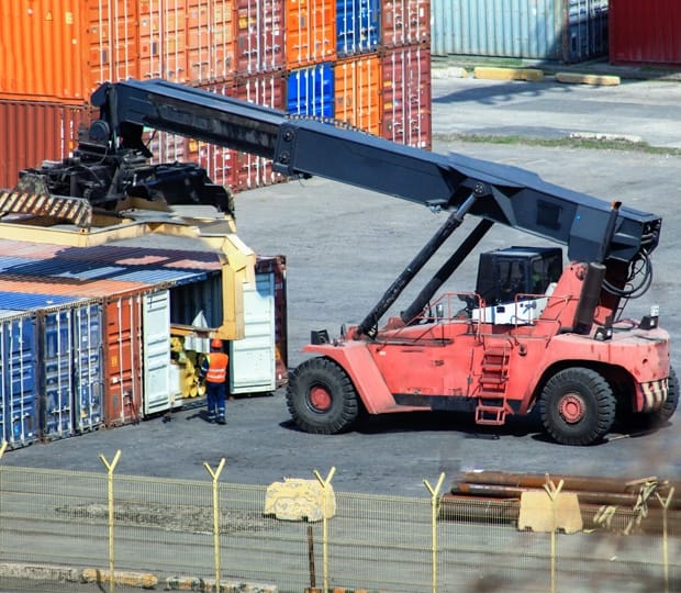 A heavy-duty crane lifting a shipping container in a port, with a worker standing nearby for supervision.