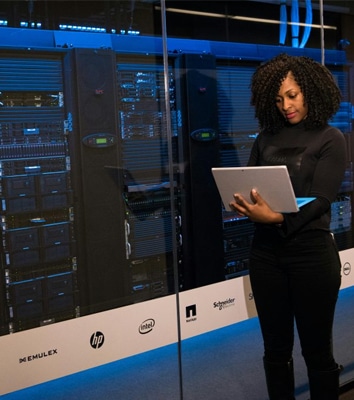 The image depicts a modern data center environment with a female IT professional or system administrator standing in front of server racks while working on a laptop or tablet.